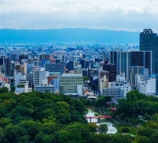 Ein Stadtbild von Osaka mit dichten modernen Gebäuden, einem großen grünen Park und vielen Bäumen im Vordergrund sowie einer kleinen roten Brücke über das Wasser. In der Ferne erheben sich Berge unter einem teilweise bewölkten Himmel, der an Ansichten in der Nähe des Kansai-Flughafens erinnert.