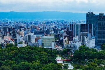 Ein Stadtbild von Osaka mit dichten modernen Gebäuden, einem großen grünen Park und vielen Bäumen im Vordergrund sowie einer kleinen roten Brücke über das Wasser. In der Ferne erheben sich Berge unter einem teilweise bewölkten Himmel, der an Ansichten in der Nähe des Kansai-Flughafens erinnert.