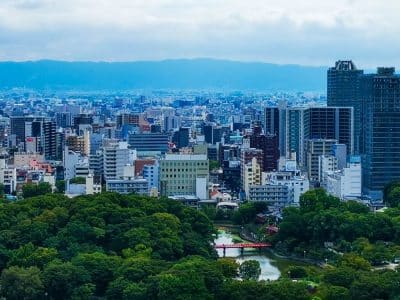 Ein Stadtbild von Osaka mit dichten modernen Gebäuden, einem großen grünen Park und vielen Bäumen im Vordergrund sowie einer kleinen roten Brücke über das Wasser. In der Ferne erheben sich Berge unter einem teilweise bewölkten Himmel, der an Ansichten in der Nähe des Kansai-Flughafens erinnert.