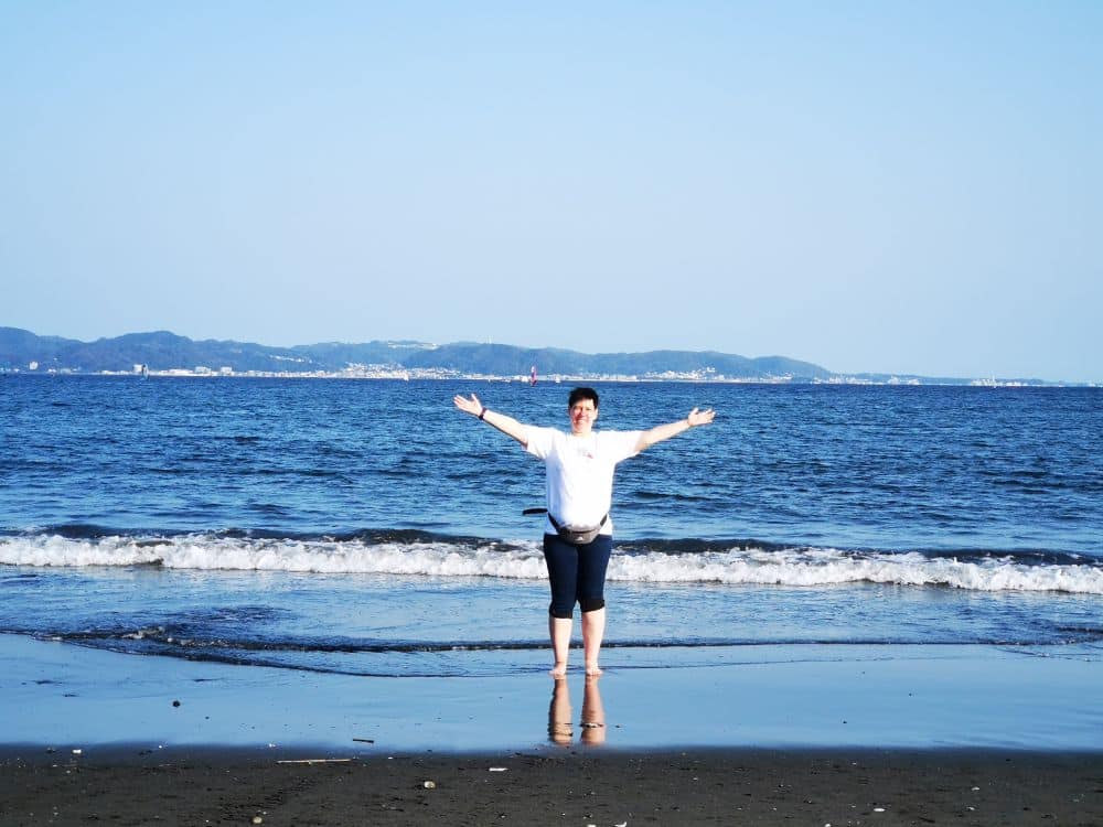 Eine Person steht mit ausgestreckten Armen auf nassem Sand in der Nähe der Küste von Enoshima und blickt in die Kamera. Im Hintergrund sind sanfte Wellen und ein ruhiges Meer zu sehen, in der Ferne Hügel unter einem klaren blauen Himmel.