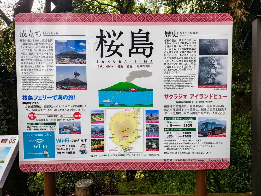 Ein Informationsschild über die Insel Sakurajima in Kagoshima mit Fotos, Karten und zweisprachigem Text über ihre Entstehung, Geschichte, Fährverbindungen und Sehenswürdigkeiten. Von der Aussichtsplattform des Shiroyama-Parks im Hintergrund sind Bäume und Grünflächen zu sehen.