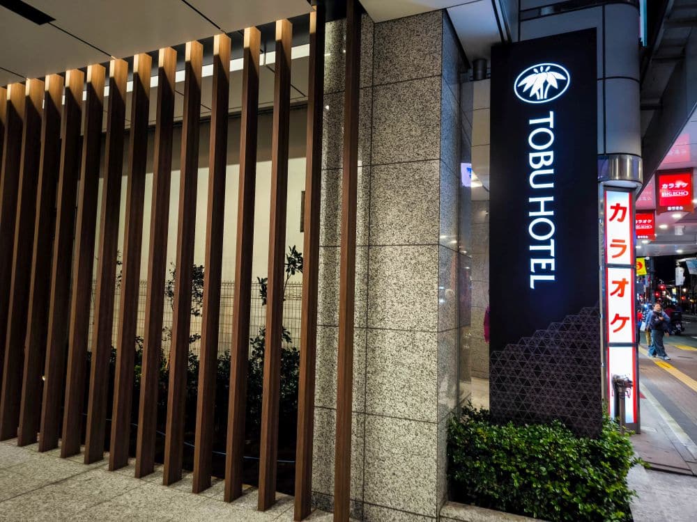 Der Eingang des Tobu Hotels ist nachts mit einem beleuchteten vertikalen Schild in Englisch und Japanisch versehen. Holzlatten und Steinmauern rahmen den Eingang ein, während die Lichter der Stadt und ein Karaoke-Schild den Bürgersteig davor beleuchten.