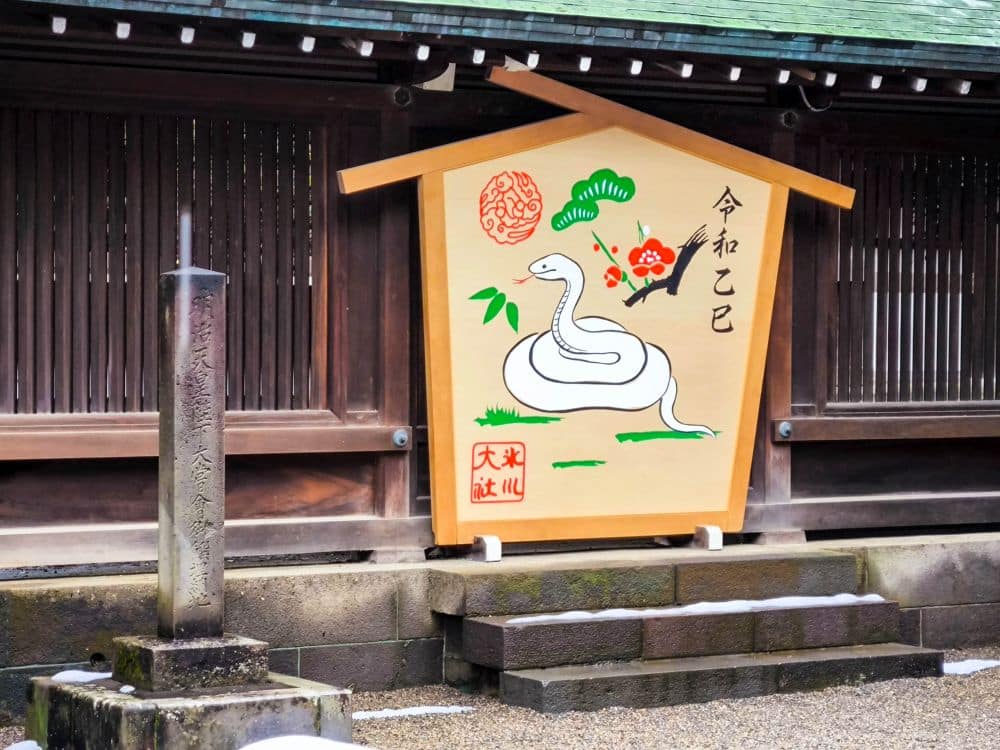 Eine Holztafel am Hikawa-Schrein zeigt eine gemalte weiße Schlange, Pflaumenblüten und japanische Schriftzeichen. Sie steht vor einer Holzstruktur mit vertikalen Latten, einer Steinsäule auf der linken Seite und Schneeflecken auf dem Boden.