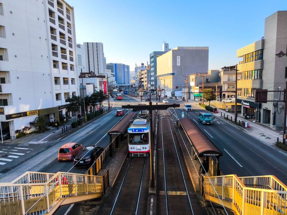 Eine rot-weiße Straßenbahn wartet an einer ebenerdigen Haltestelle im Herzen des Tipps Nagasaki, umgeben von Gebäuden, Autos und Fußgängerwegen unter einem klaren blauen Himmel.