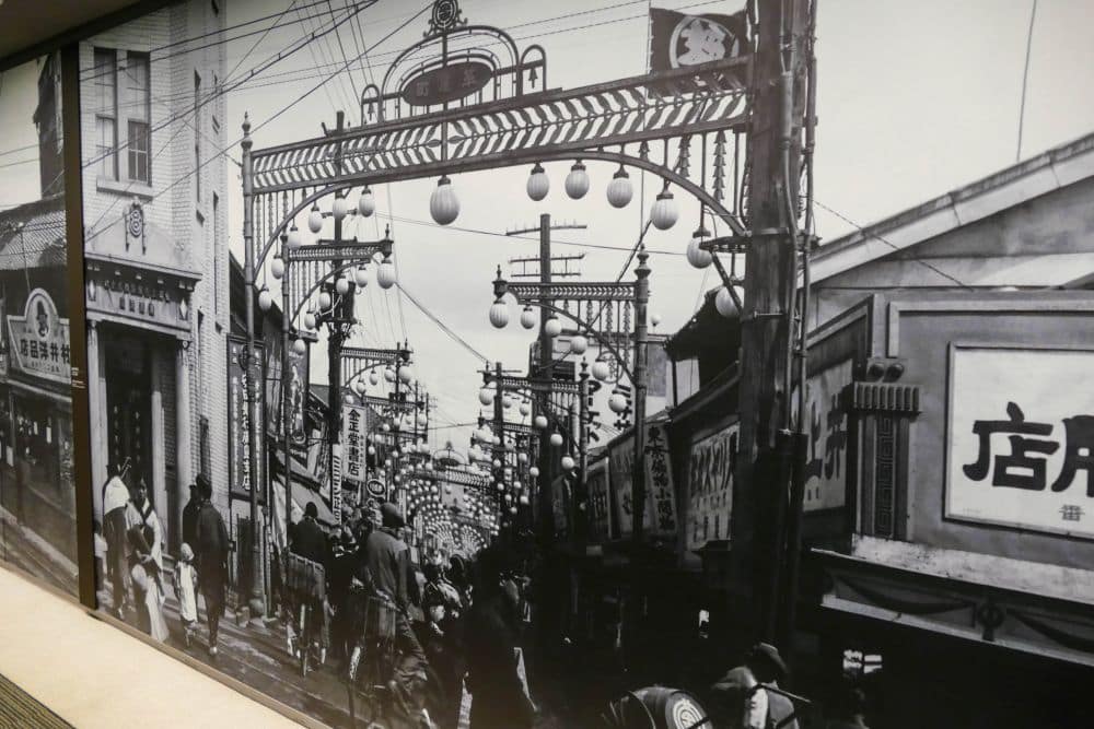 Schwarz-Weiß-Foto einer belebten japanischen Straße, die von Geschäften und Schildern gesäumt ist und in der viele Menschen unterwegs sind. Über der Straße kreuzen Laternen und Stromleitungen die Straße und verbinden lebendige Energie mit einem Gefühl von historischem Frieden.