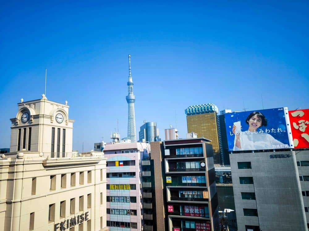 Ein Stadtbild in Tokio mit dem ikonischen Tokyo Skytree, modernen Tobu-Gebäuden, einem Uhrenturm und einer großen Werbetafel, die eine lächelnde Person mit einem Getränk in der Hand vor einem klaren blauen Himmel zeigt.