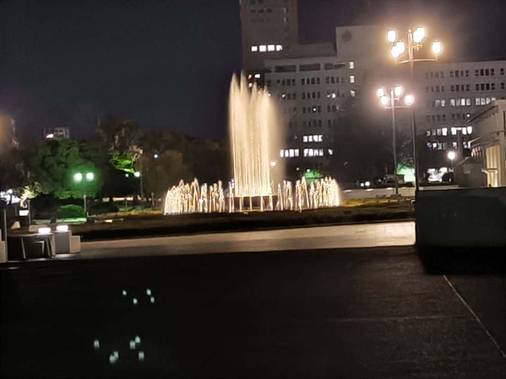 Ein hell erleuchteter Springbrunnen spritzt nachts in einem Stadtpark Wasser in die Höhe und strahlt zwischen leuchtenden Straßenlaternen und weit entfernten, beleuchteten Gebäuden im Hintergrund Ruhe aus.