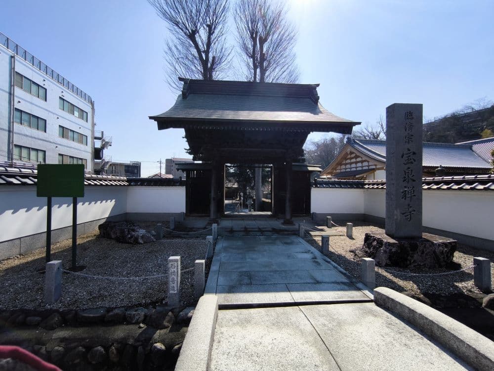 Ein traditionelles japanisches Tempeltor am Hosenji-Tempel in Hino, durch das ein steinerner Weg führt, flankiert von weißen Mauern und einem hohen, mit japanischen Schriftzeichen beschrifteten Denkmal, das die reiche Geschichte unter einem klaren blauen Himmel widerspiegelt.