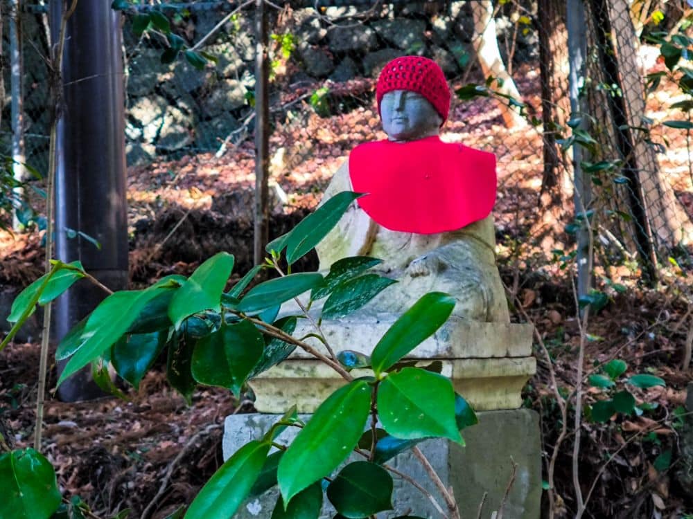 Eine verwitterte Steinstatue einer sitzenden Figur mit roter Strickmütze und Lätzchen sitzt in Takao zwischen grünen Blättern, eingebettet in ein bewaldetes Außengelände mit einem Drahtzaun und einer Steinmauer dahinter.