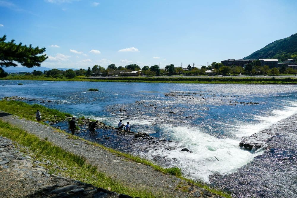 Eine malerische Flusslandschaft in Arashiyama mit klarem Wasser, das über Felsen fließt, ein paar Menschen, die am Ufer stehen, und Bäumen und Hügeln im Hintergrund unter strahlend blauem Himmel.