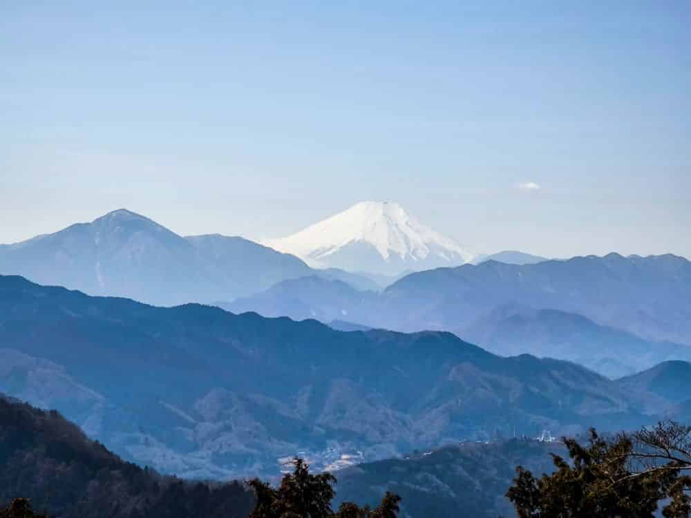 Der schneebedeckte Berg Fuji erhebt sich in der Ferne hinter blauen und violetten Gebirgsketten, darunter auch Takao, unter einem klaren Himmel, mit einer Andeutung von Baumkronen am unteren Bildrand.