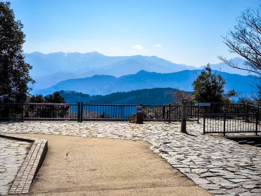 Ein malerischer Aussichtspunkt in Takao mit einem gepflasterten Steinweg und einem Geländer bietet einen Blick auf ferne, blaue Berge unter einem klaren Himmel. Bäume umrahmen die ruhige Szene auf beiden Seiten.