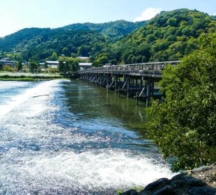 Eine Holzbrücke führt über einen klaren Fluss mit sanften Stromschnellen und üppig grünen Bäumen, der sich vor dem Hintergrund der bewaldeten Hügel von Arashiyama unter einem strahlend blauen Himmel erstreckt.