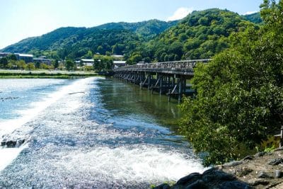 Eine Holzbrücke führt über einen klaren Fluss mit sanften Stromschnellen und üppig grünen Bäumen, der sich vor dem Hintergrund der bewaldeten Hügel von Arashiyama unter einem strahlend blauen Himmel erstreckt.