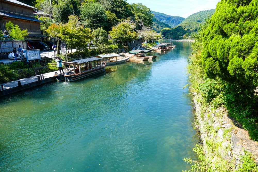 Ein klarer Fluss fließt durch Arashiyama, an dessen Ufer traditionelle Holzboote angedockt sind, umgeben von üppig grünen Bäumen und Hügeln unter einem strahlend blauen Himmel, und auf der linken Seite sind Holzhäuser zu sehen.