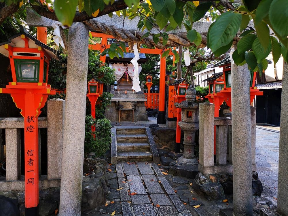 Ein kleiner Shinto-Schrein mit roten Torii-Toren und Steinlaternen liegt friedlich unter dem Mond, sein steinerner Weg ist von grünem Laub und verstreutem Laub umgeben. Der ruhige Bereich ist von Bäumen beschattet und lädt zum Nachdenken ein.