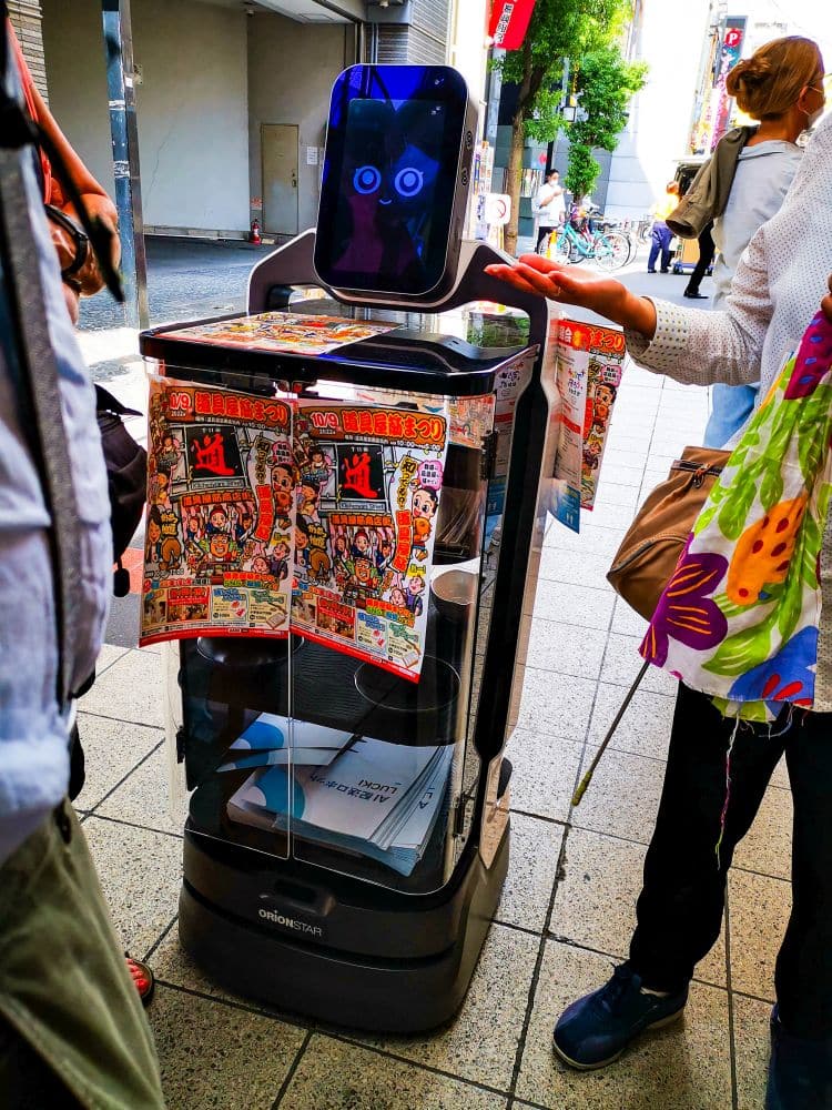 Ein Lieferroboter mit digitalem Gesicht und ausdrucksstarken Augen steht auf einem städtischen Gehweg, der mit bunten Veranstaltungsflyern geschmückt ist. Als freundlicher Fremdenführer interagiert der Roboter mit den Menschen in der Nähe, während im Hintergrund Fahrräder vorbeifahren.