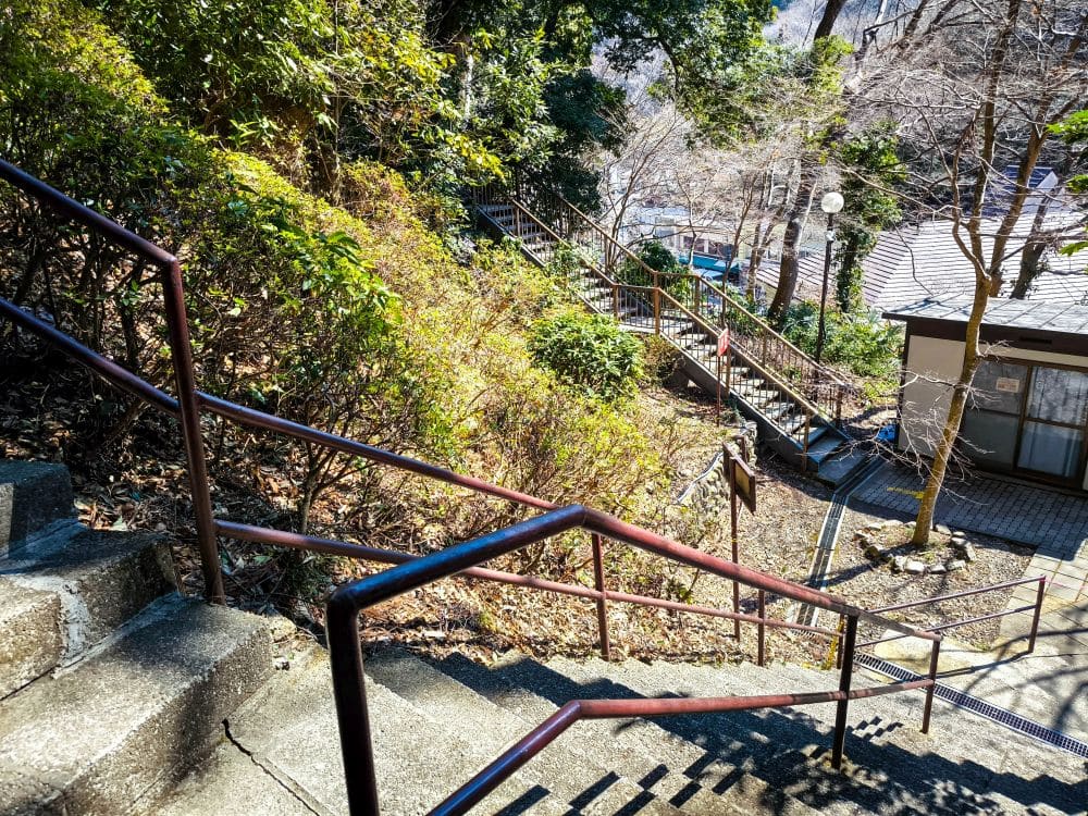 Eine Betontreppe mit Metallgeländer schlängelt sich an einem von grünen Büschen und Bäumen umgebenen Hang in Takao hinunter. Das Sonnenlicht fällt durch die Äste, und unten rechts ist ein Gebäude teilweise sichtbar.