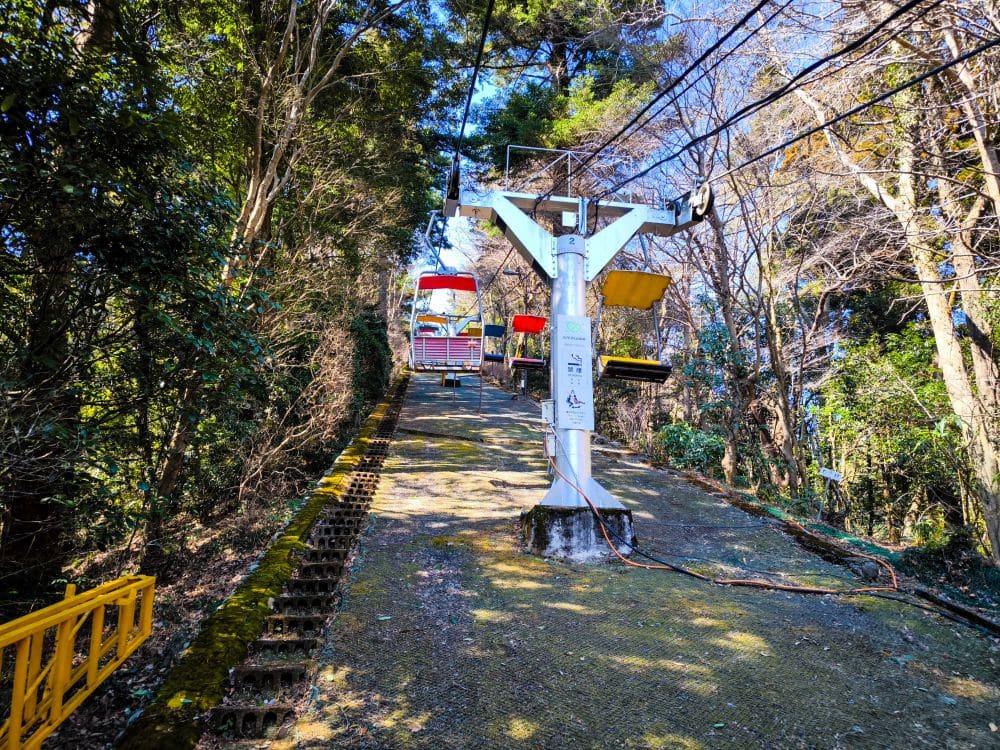 Ein Sessellift mit roten und gelben Sitzen führt über einen steilen, moosbewachsenen Takao-Weg, der auf beiden Seiten von Bäumen umgeben ist, unter einem klaren blauen Himmel nach oben. Auf der linken Seite verläuft parallel zum Lift eine Treppe.