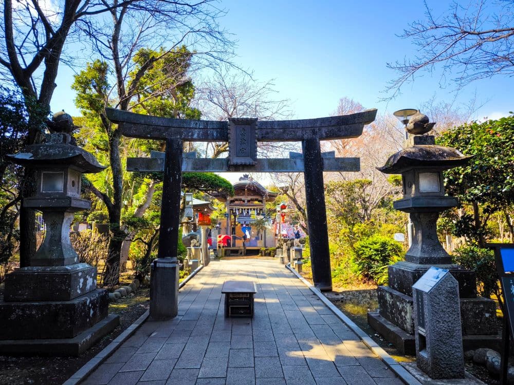 Das steinerne Torii-Tor am Eingang zum Okutsunomiya, flankiert von zwei Steinlaternen, mit einem Weg, der zu dem farbenfrohen Schreingebäude führt, umgeben von Bäumen und Grün an einem sonnigen Tag.