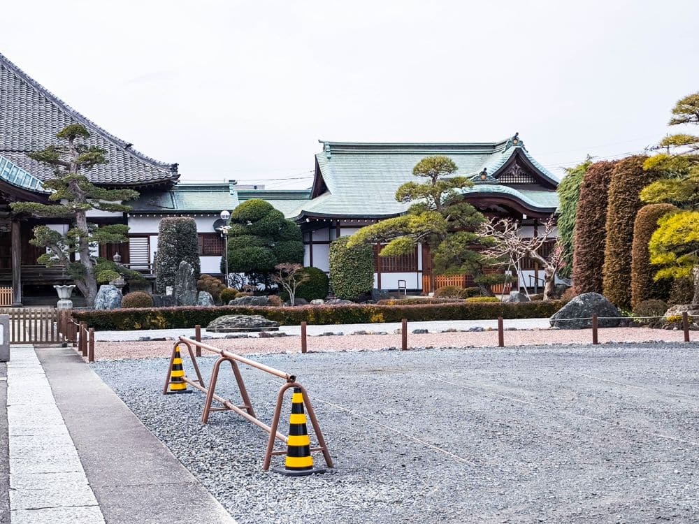 Ein traditionelles japanisches Gebäude in Kawagoe mit grünen Ziegeldächern, ordentlich beschnittenen Bäumen, Kiesboden und zwei gelb-schwarzen Verkehrskegeln vor einer niedrigen Metallschranke.