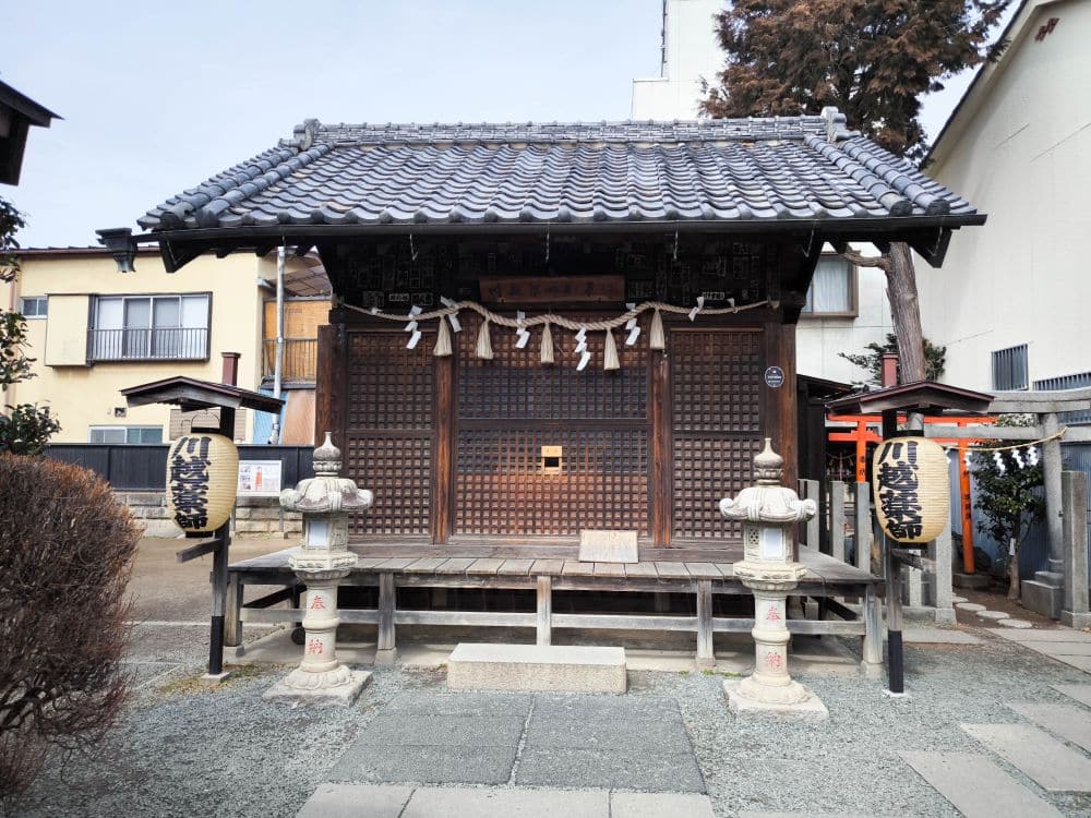 Ein kleiner traditioneller japanischer Schrein in Kawagoe mit einem Ziegeldach, Luftschlangen, Laternen und Holzstufen, umgeben von Gebäuden und Bäumen an einem klaren Tag.