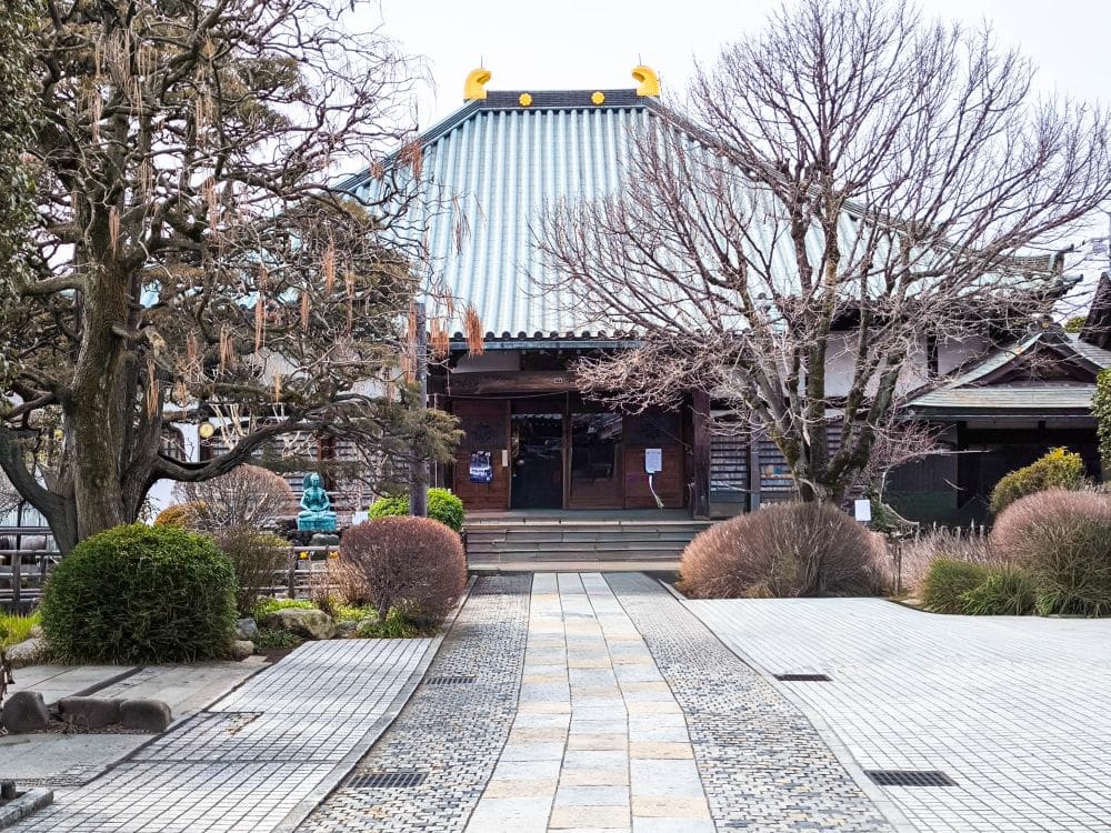 Ein traditioneller japanischer Tempel in Kawagoe mit einem grünen Ziegeldach, umgeben von kahlen Bäumen und gepflegten Sträuchern, steht an einem bewölkten Tag am Ende eines steinernen Weges.