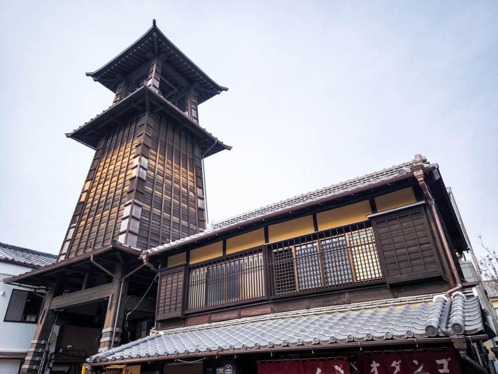 Der ikonische Uhrenturm Toki no Kane steht neben einem Gebäude im alten Stil mit Ziegeldächern und hölzernen Gitterfenstern und fängt den zeitlosen Charme des historischen Kawagoe vor einem blassen Himmel ein.