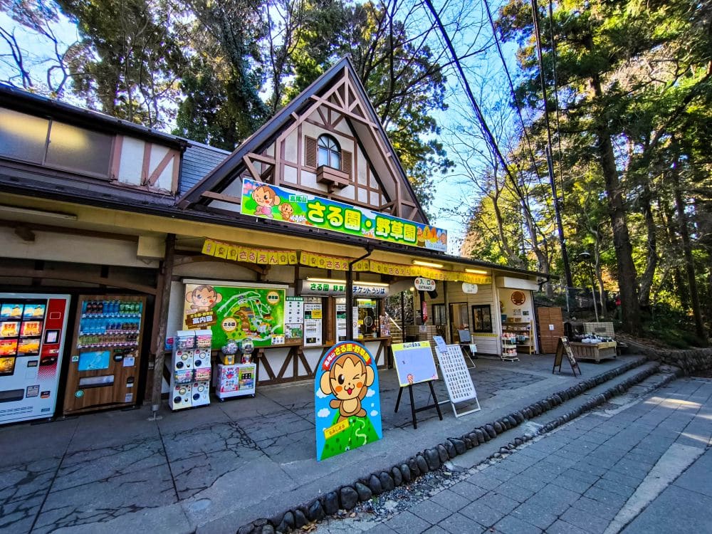 Ein charmantes Geschäft in der Nähe von Takao mit einem dreieckigen Dach, bunten Schildern mit Cartoon-Affen, Automaten und verschiedenen Auslagen, umgeben von hohen Bäumen an einem sonnigen Tag.