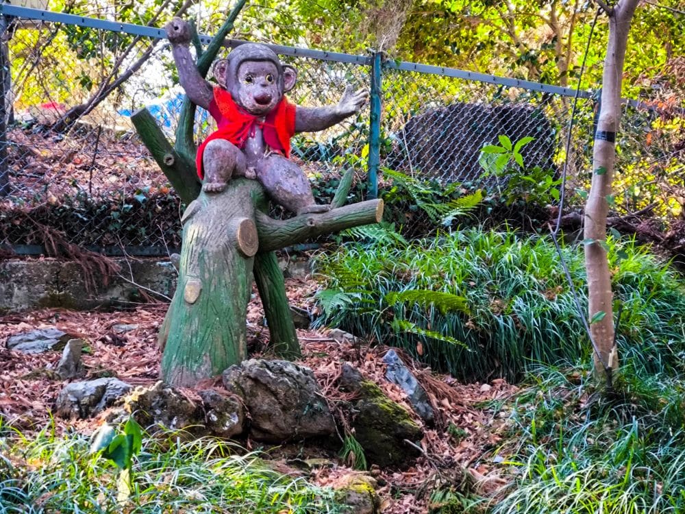 Die Statue eines Affen mit roter Weste sitzt auf einem Baumstumpf in der bewaldeten Gegend von Takao in der Nähe eines Drahtzauns, umgeben von Steinen, Blättern und üppigem Grün.