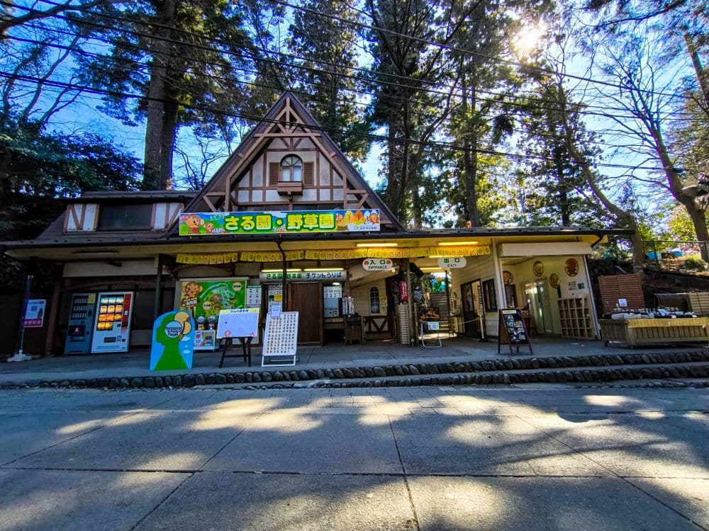 Ein malerisches Gebäude mit einem steilen, dreieckigen Dach und Schildern in japanischer Sprache steht zwischen hohen Bäumen in der Nähe von Takao. An einem sonnigen Tag sind in der Nähe des Eingangs mehrere Verkaufsautomaten und Informationstafeln aufgestellt.