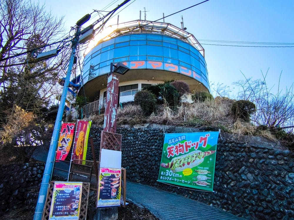 Ein rundes, gläsernes Gebäude steht auf einem Hügel in Takao. Japanische Schilder und farbenfrohe Banner werben für Lebensmittel entlang eines abschüssigen Steinwegs, der zu ihm hinaufführt. Bäume und ein blauer Himmel umgeben die lebhafte Szene.