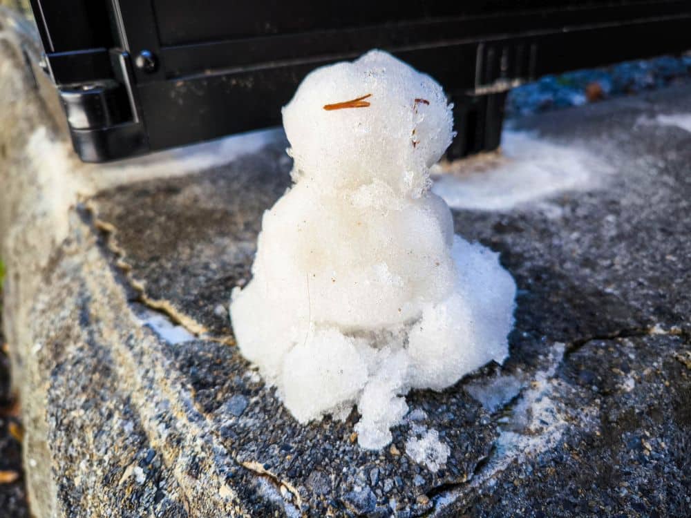 Ein kleiner, grob geformter Schneemann mit einem Stock als Nase sitzt auf einer Betonfläche in der Nähe eines schwarzen Objekts in Takao, das mit Schnee und Eis bedeckt ist.
