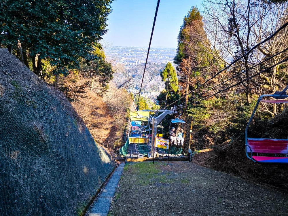 Die Menschen fahren mit bunten, offenen Sesselliften die steilen, von Bäumen gesäumten Hänge von Takao hinauf, während in der Ferne unter klarem Himmel eine Stadtlandschaft zu sehen ist.