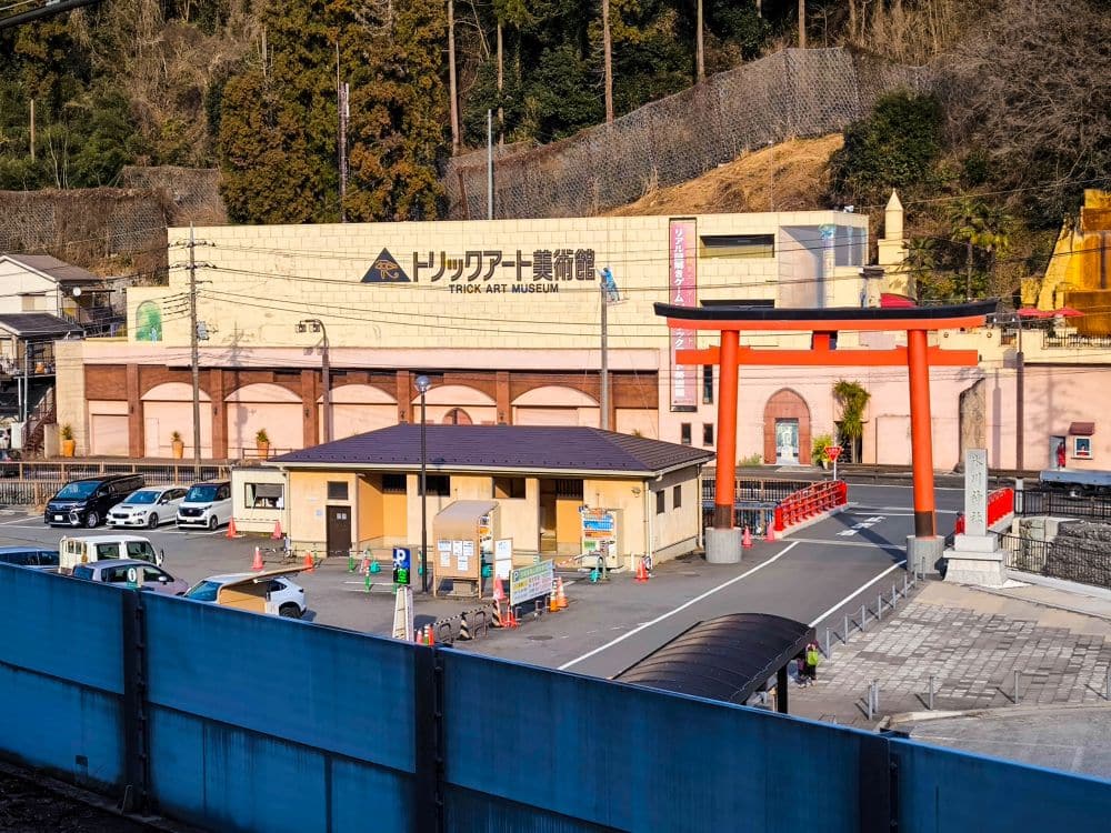 Ein Parkplatz mit mehreren Autos und orangefarbenen Kegeln befindet sich vor einem Gebäude mit der Aufschrift Trick Art Museum in der Nähe von Takao. Ein großes rotes Torii-Tor und eine kleine Brücke sind ebenfalls zu sehen, mit bewaldeten Hügeln im Hintergrund.