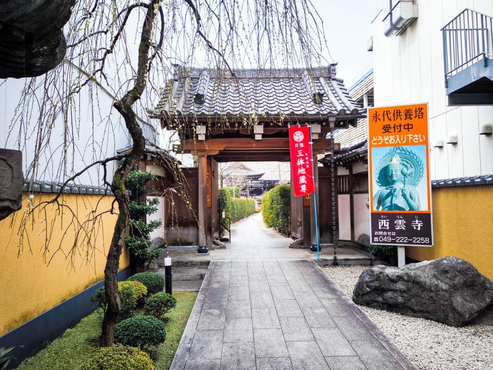 Ein traditioneller japanischer Tempeleingang in Kawagoe mit einem Holztor, gelben Wänden, gepflegten Büschen und Bannern mit japanischem Text. Neben dem Weg sind ein Statuenplakat und ein Baum mit kahlen Ästen zu sehen.