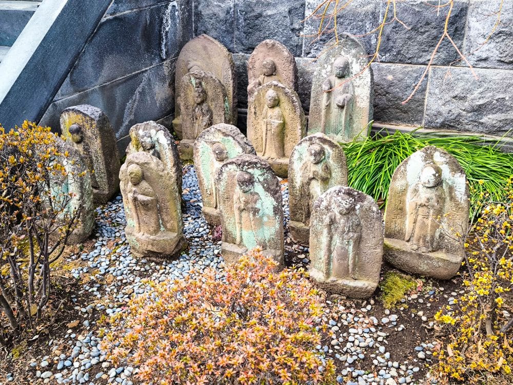 Mehrere verwitterte Steinstatuen, die buddhistischen Figuren ähneln, sind in einem kleinen Gartenbereich mit Kies, Büschen und Gras vor einer Steinmauer und einer Treppe in Kawagoe aufgestellt.