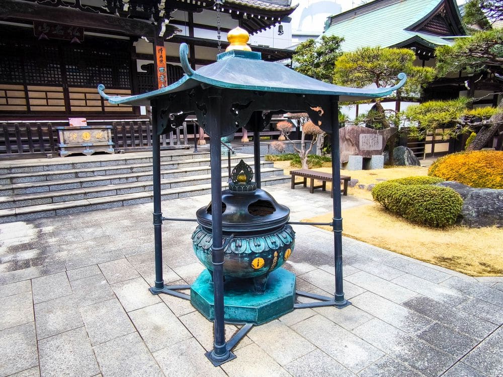 Großes Räuchergefäß aus Metall mit einem dekorativen Dach, aufgestellt auf einem Steinhof vor einem traditionellen Kawagoe-Tempelgebäude, umgeben von gepflegten Sträuchern und Steinstufen.