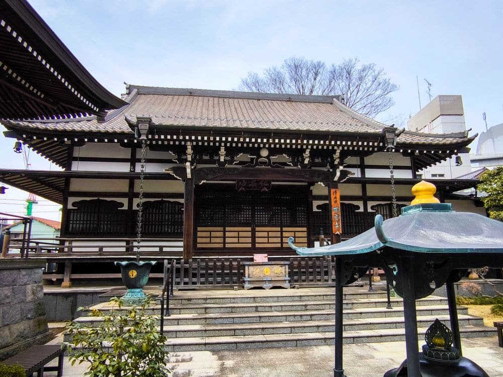 Ein traditioneller japanischer Tempel in Kawagoe mit einem Ziegeldach, einer Holzkonstruktion, Steinstufen, die zum Eingang führen, und einer dekorativen Laterne im Vordergrund. Im Hintergrund sind Bäume und moderne Gebäude zu sehen.