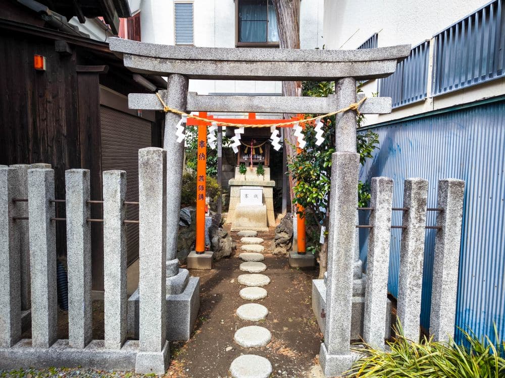 Ein kleiner Shinto-Schrein in Kawagoe, eingebettet zwischen Gebäuden, verfügt über ein steinernes Torii-Tor, ein rotes Holztor, Steinlaternen und runde Trittsteine, die zum Altar führen - ein Echo des zeitlosen Geistes von Toki no Kane und klingendem Herz.