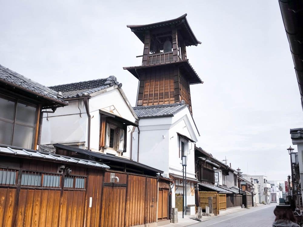 Traditionelle japanische Straße in Kawagoe mit hölzernen und weiß verputzten Gebäuden, mit dem ikonischen Uhrenturm Toki no Kane unter einem bewölkten Himmel. Die ruhige Straße ist leer, was ein Gefühl der Zeitlosigkeit hervorruft.