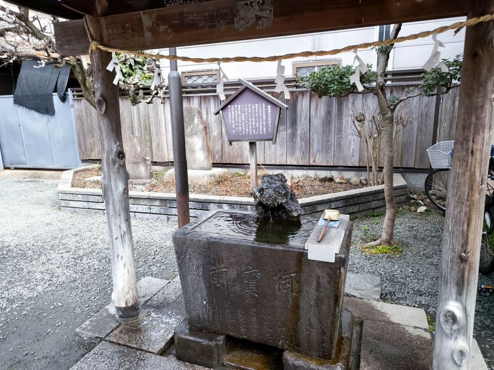 Ein traditioneller japanischer Reinigungsbrunnen mit einem Steinbecken, einer Schöpfkelle und einem hölzernen Unterstand in einem kleinen Außenbereich in der Nähe des Toki no Kane in Kawagoe, eingerahmt von Kies und einem Holzzaun im Hintergrund.