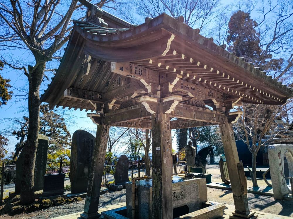 Ein traditionelles japanisches Tempeltor aus Holz mit einem schrägen Dach steht zwischen Grabsteinen und Bäumen unter dem klaren blauen Himmel von Takao. Das Sonnenlicht fällt durch die Äste und wirft Schatten auf die Steinstrukturen darunter.