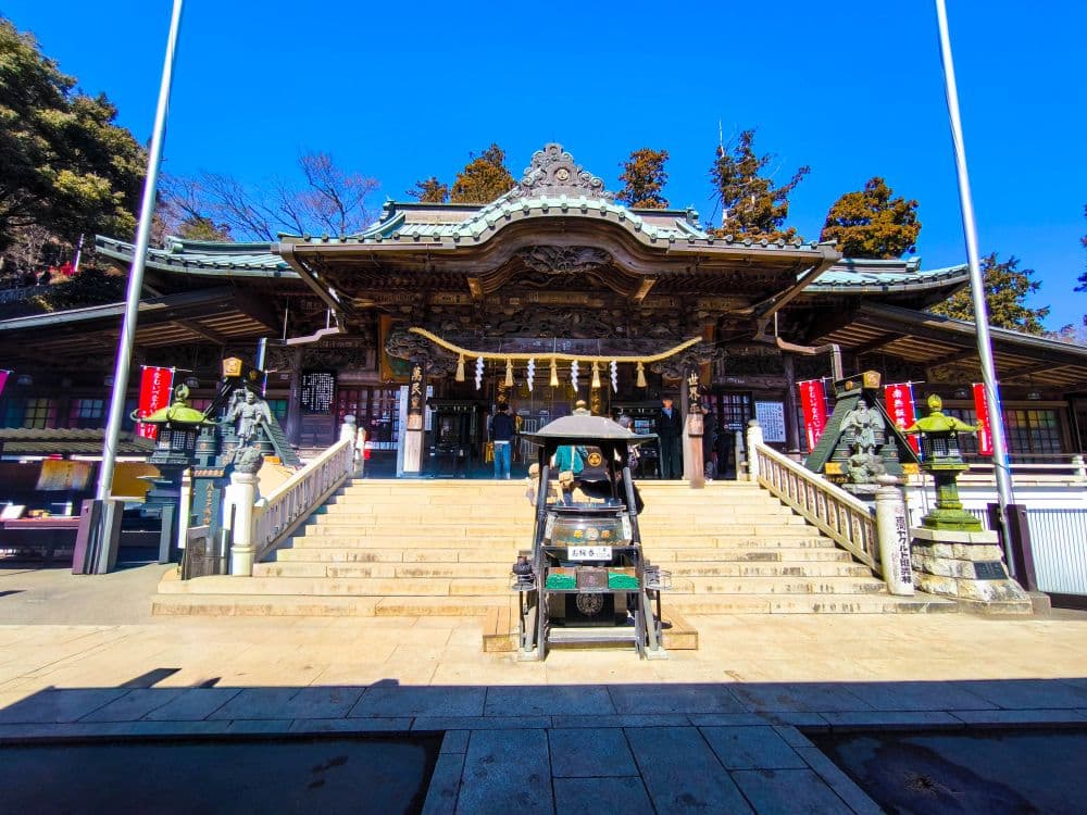 Traditioneller japanischer Takao-Schrein mit verschnörkeltem Dach, Steinlaternen und dekorativen Elementen. Eine Treppe führt zum Eingang, der von Wächterstatuen flankiert wird. In der Mitte der Treppe ist bei hellem Tageslicht eine Rikscha geparkt.
