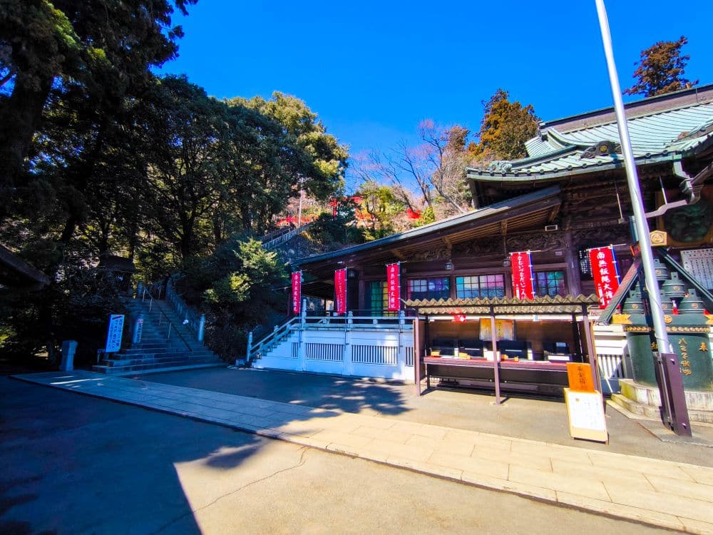 Ein traditioneller japanischer Tempel mit grünen Ziegeldächern steht neben Steinstufen auf dem Berg Takao, umgeben von üppigen Bäumen unter einem strahlend blauen Himmel. Rote Banner mit weißer Schrift hängen an der Außenseite des Gebäudes.