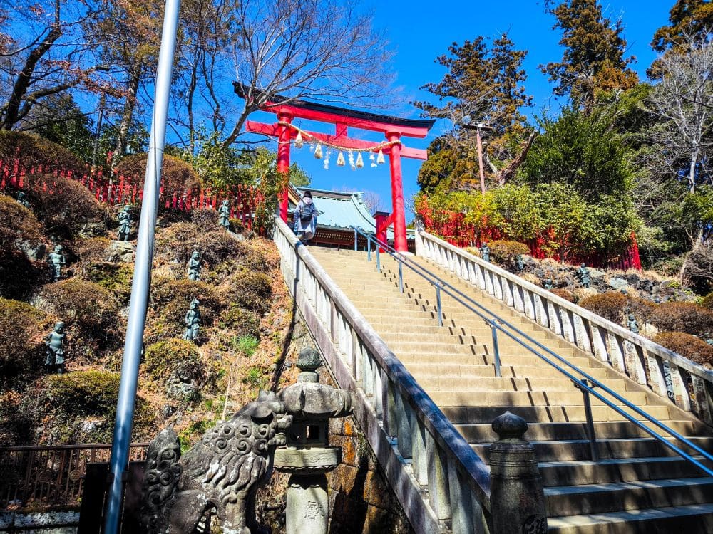 Eine lange Steintreppe in Takao führt hinauf zu einem leuchtend roten Torii-Tor an einem japanischen Schrein, umgeben von üppigem Grün und Steinstatuen unter einem klaren blauen Himmel.