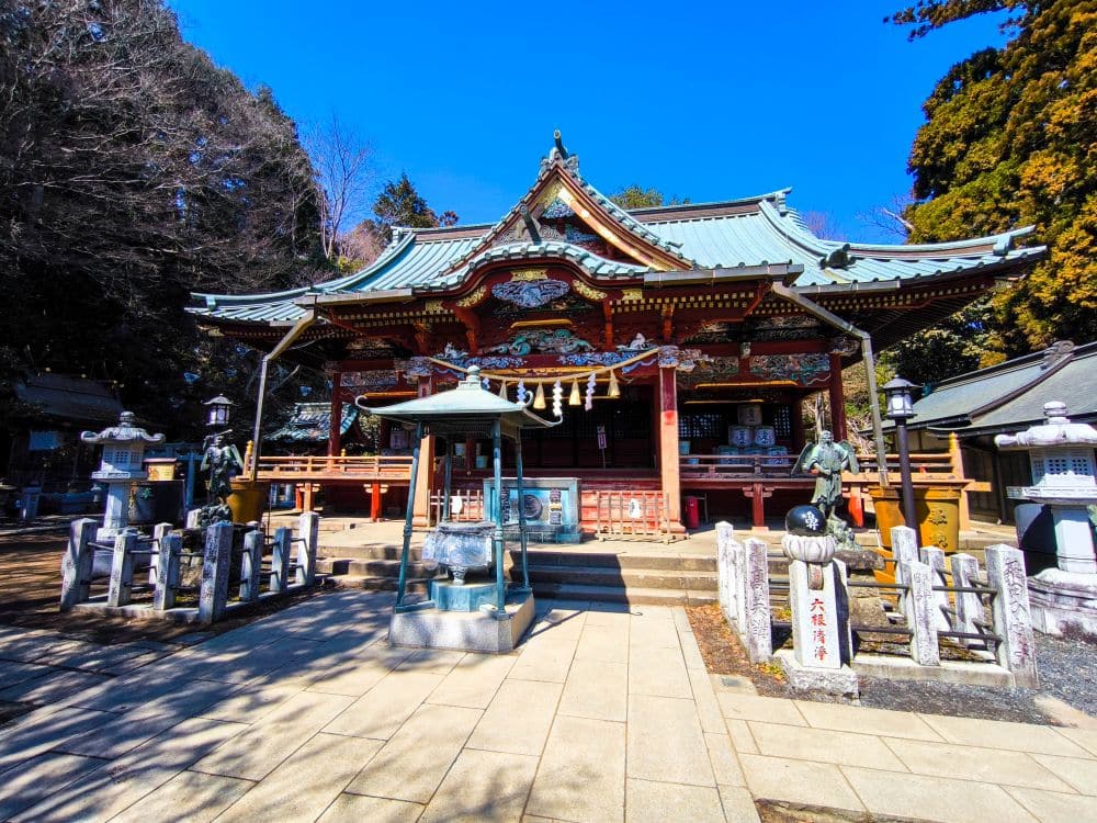Ein traditioneller japanischer Schrein auf Takao mit kunstvollen Holzschnitzereien, einem grünen Ziegeldach und dekorativen Details. Davor stehen Steinlaternen und Statuen, umgeben von hohen Bäumen unter einem strahlend blauen Himmel.