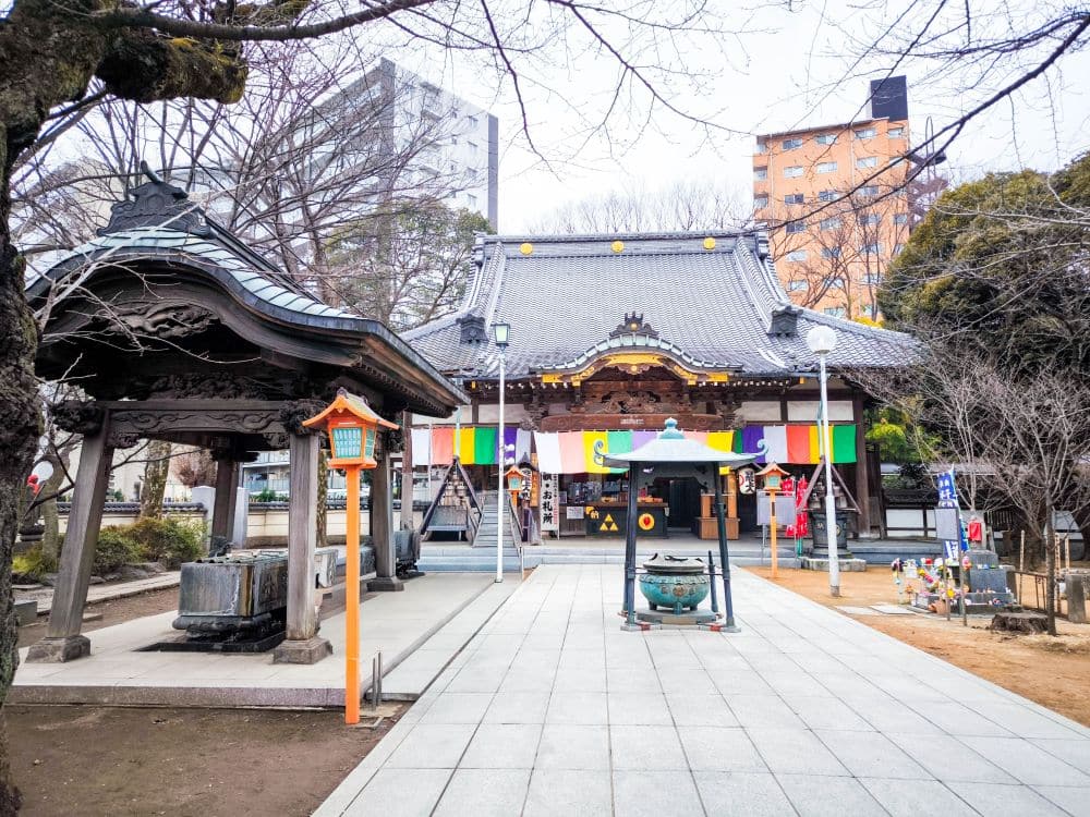Ein japanischer Tempelhof in Kawagoe mit einer mit bunten Bannern geschmückten Haupthalle, einem Glockenpavillon auf der linken Seite und einem großen Räuchergefäß in der Mitte. Laublose Bäume und moderne Gebäude umgeben den ruhigen Tempel.