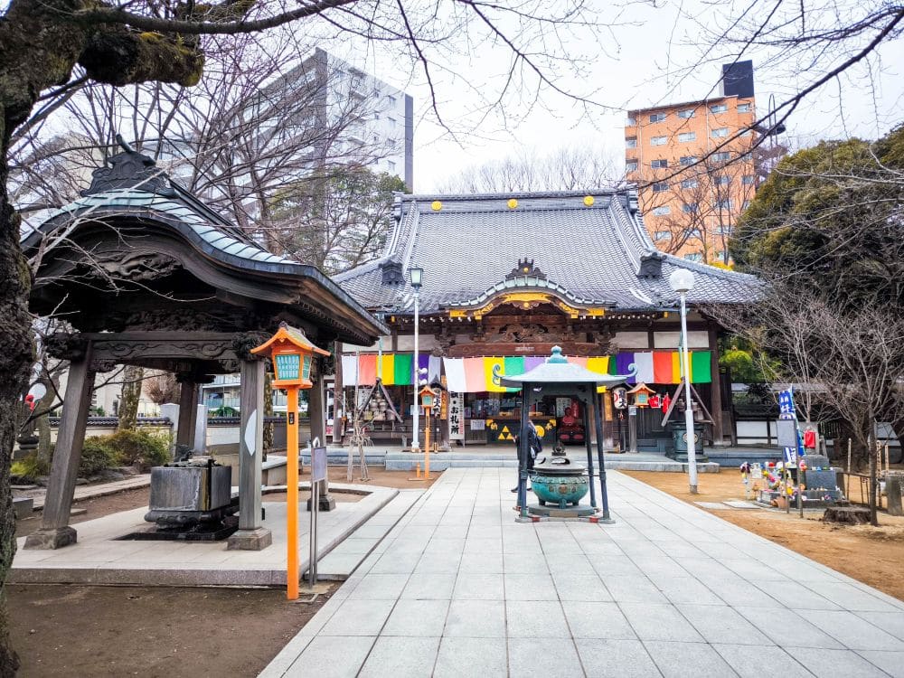 Ein japanischer Tempel mit bunten Bannern steht im Zentrum von Kawagoe, flankiert von einem Glockenpavillon auf der linken Seite und städtischen Gebäuden im Hintergrund. Laublose Bäume säumen den Steinweg, der zum Tempeleingang führt.