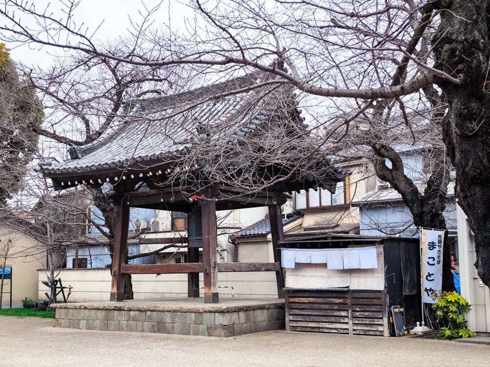 Ein traditioneller japanischer Glockenpavillon mit Ziegeldach steht inmitten kahler Bäume in einem ruhigen Hof in Kawagoe, neben Holzgebäuden und einem Schild mit japanischer Schrift.